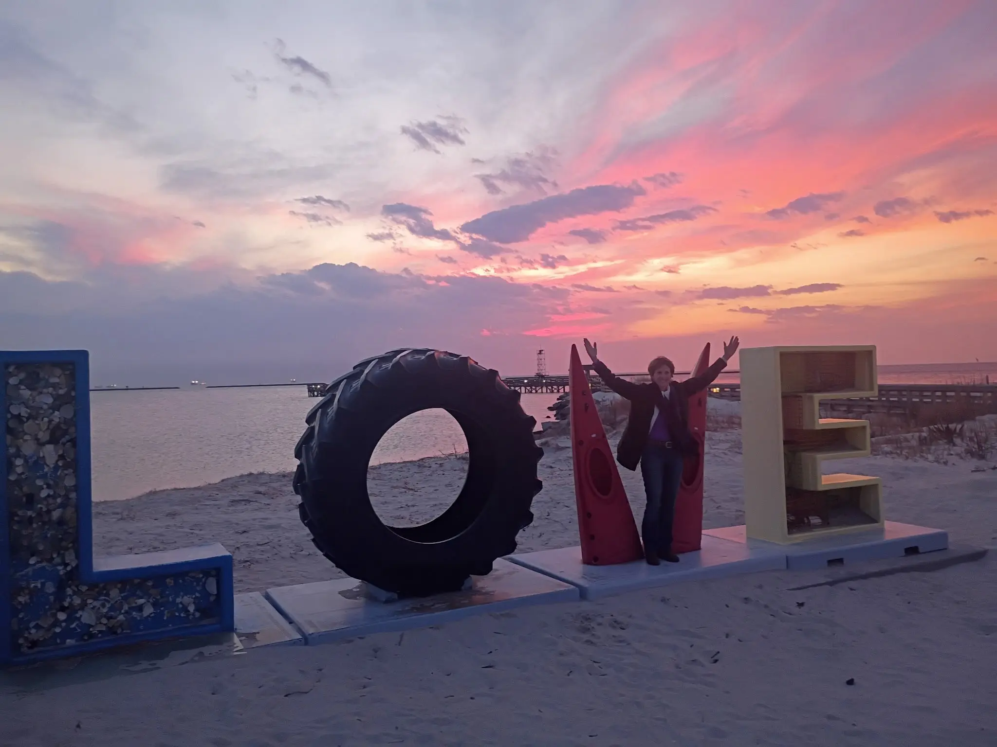 Woman posing at the Cape Charles LOVE sign on the beach at sunset with the Chesapeake Bay and pier in the background