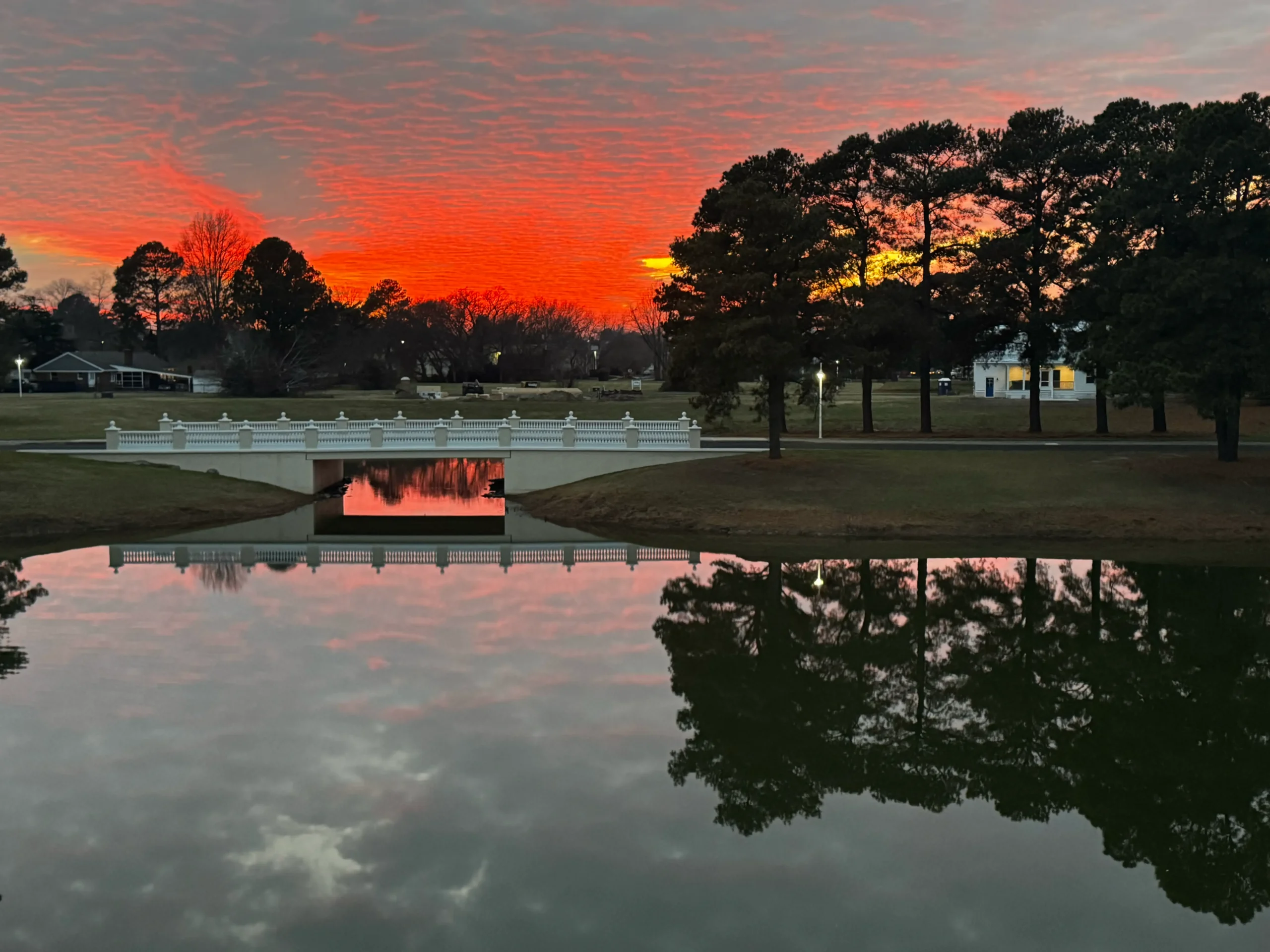 A fiery red and orange sunset reflected in the still water of Central Park pond in Cape Charles, Virginia, with the iconic white bridge and pine trees silhouetted against the evening sky.A fiery red and orange sunset reflected in the still water of Central Park pond in Cape Charles, Virginia, with the iconic white bridge and pine trees silhouetted against the evening sky.