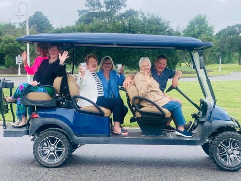 Group of friends riding and waving from a blue six-seater golf cart in Cape Charles, showing why golf carts in Cape Charles are a favorite way to get around